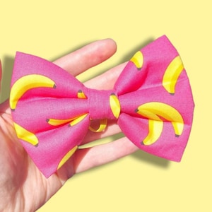 May include: A pink bow tie with a yellow banana pattern. The bow tie is held in a hand.