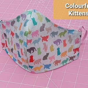 May include: A white face mask with a colorful kitten print. The mask has pink elastic straps and is on a pink cutting mat.