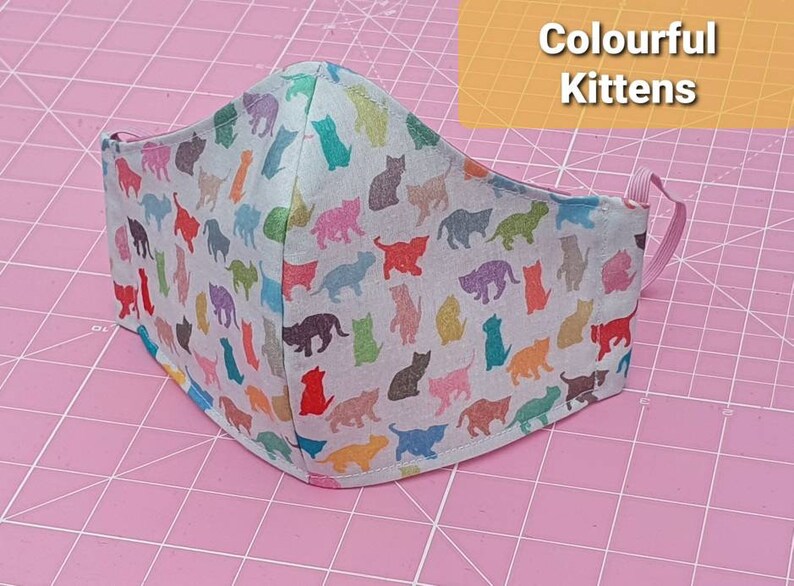 May include: A white face mask with a colorful kitten print. The mask has pink elastic straps and is on a pink cutting mat.