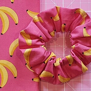 Puede incluir: Un scrunchie de tela rosa con estampado de pl&aacute;tanos amarillos. El scrunchie est&aacute; hecho de una tela suave y el&aacute;stica, ideal para sujetar el cabello.
