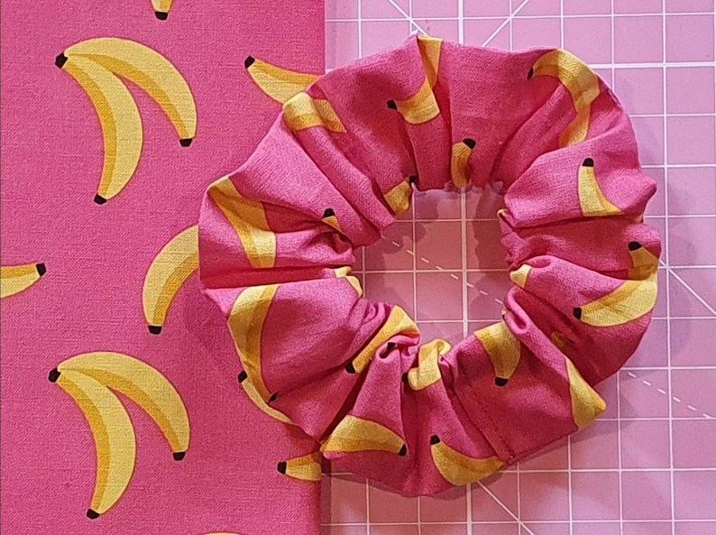 Puede incluir: Un scrunchie de tela rosa con estampado de pl&aacute;tanos amarillos. El scrunchie est&aacute; hecho de una tela suave y el&aacute;stica, ideal para sujetar el cabello.