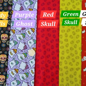 May include: Five fabric swatches featuring different Halloween designs. The first swatch is black with white candy skulls and flowers. The second swatch is purple with white ghosts wearing rainbow hats. The third swatch is red with white skulls. The fourth swatch is yellow with green skulls. The fifth swatch is red with white stars and white ghosts.