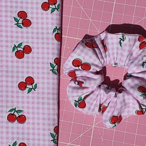 Könnte beinhalten: Ein rosa Gingham-Stoff-Scrunchie mit Kirschendruck. Das Scrunchie ist aus weichem, dehnbarem Stoff und perfekt, um Haare zurückzuhalten.