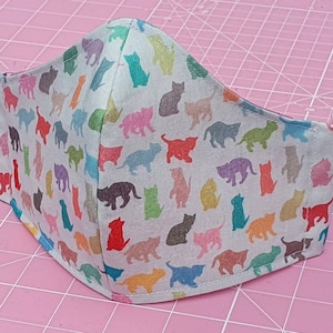 May include: A white face mask with a colorful cat print. The mask is made of fabric and has a pink elastic band.