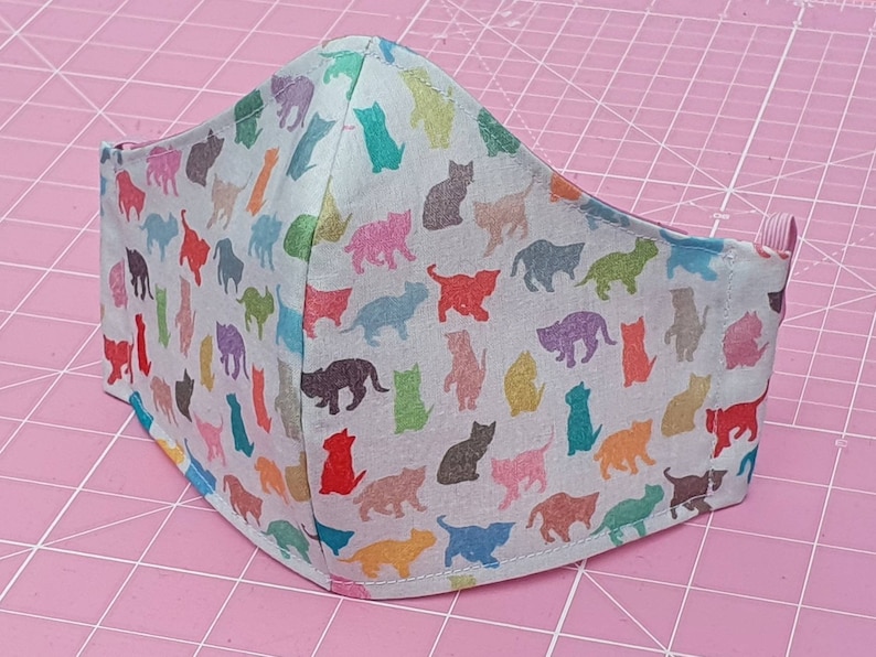 May include: A white face mask with a colorful cat print. The mask is made of fabric and has a pink elastic band.