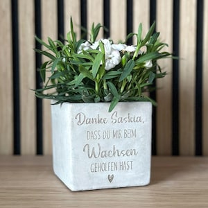 Könnte beinhalten: Ein quadratischer, hellgrauer Beton-Pflanztopf mit dem Text "Danke Saskia, DASS DU MIR BEIM Wachsen GEHOLFEN HAST" und einem Herzsymbol. Der Topf enthält grüne Pflanzen mit weißen Blüten. Der Hintergrund ist eine Holzwand mit schwarzen Streifen.