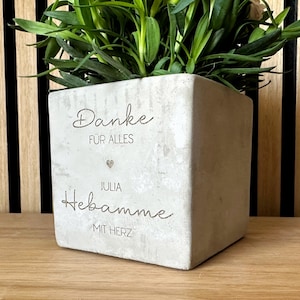 Könnte beinhalten: Ein quadratischer, hellgrauer Betonblumentopf mit den Worten "Danke für alles" und "Julia Hebamme mit Herz" auf der Vorderseite eingraviert. Der Topf enthält grüne Blätterpflanzen. Der Hintergrund ist eine Holzoberfläche und eine vertikal gestreifte Wand.