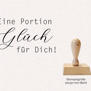 Könnte beinhalten: Ein Holzstempel mit hellem Griff und rechteckiger Basis. Der Stempel trägt den deutschen Text "Eine Portion Glück für Dich!" und die Maße "20x30 mm (BxH)".