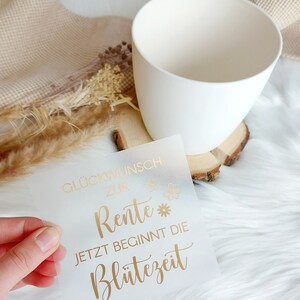 May include: A white card with gold lettering that reads "Gl&uuml;ckwunsch zur Rente* Jetzt beginnt die Bl&uuml;tezeit".