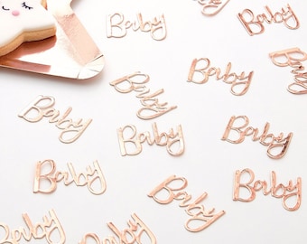 Può includere: Confetti color oro rosa con la parola "Baby" scritta su ogni pezzo. I confetti sono sparsi su una superficie bianca.