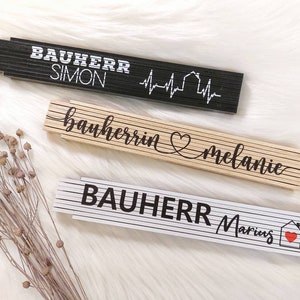 Könnte beinhalten: Drei Holz-Klapplineale mit schwarzem und weißem Text. Das obere Lineal sagt "Bauherr Simon" mit einem Herz und einer Hausgrafik. Das mittlere Lineal sagt "bauherrin melanie" mit einem Herz. Das untere Lineal sagt "Bauherr Marius" mit einem Herz und einer Hausgrafik.