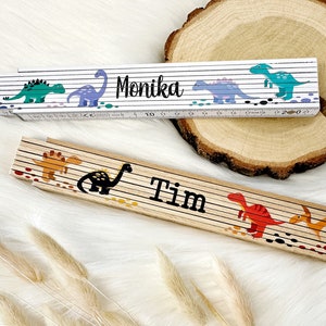 Könnte beinhalten: Zwei Holz-Klapplineale mit Dinosaurier-Illustrationen und personalisierten Namen. Das obere Lineal ist weiß mit blauen, grünen und lila Dinosauriern und dem Namen "Monika". Das untere Lineal ist braun mit orangefarbenen, roten und schwarzen Dinosauriern und dem Namen "Tim".