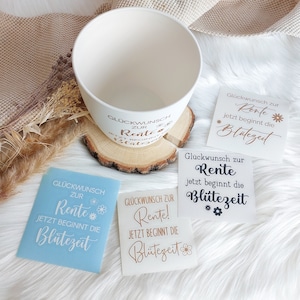May include: Three white cards with the text "Gl&uuml;ckwunsch zur Rente! Jetzt beginnt die Bl&uuml;tezeit" in gold lettering. The cards are on a white fluffy surface with a white ceramic bowl and a wooden base.