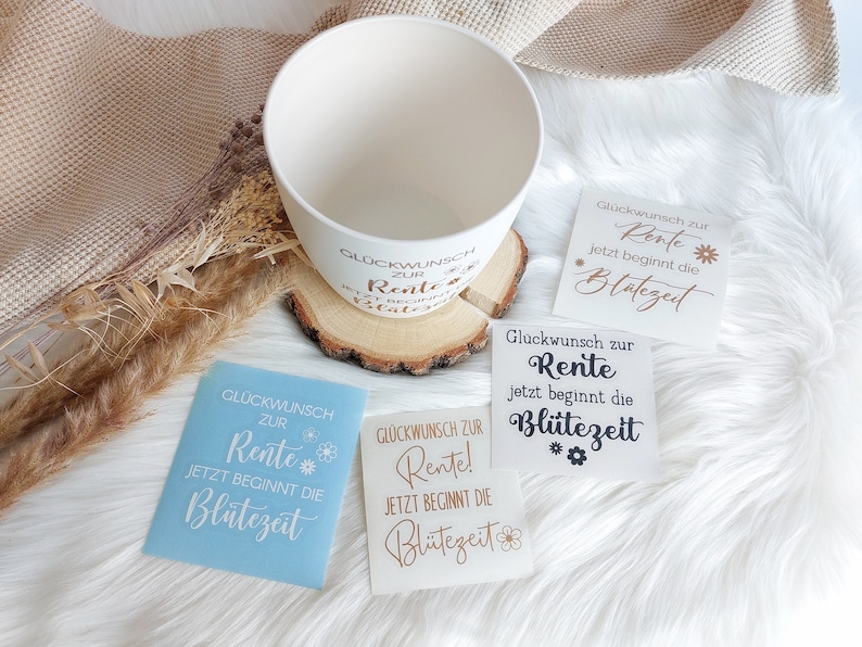 May include: Three white cards with the text "Gl&uuml;ckwunsch zur Rente! Jetzt beginnt die Bl&uuml;tezeit" in gold lettering. The cards are on a white fluffy surface with a white ceramic bowl and a wooden base.