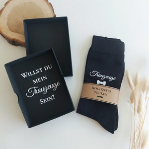 Könnte beinhalten: Eine schwarze Geschenkbox mit dem weißen Text "WILLST DU MEIN Trauzeuge SEIN?" neben einem Paar schwarzer Socken mit dem Wort "Trauzeuge" und einer Fliege. Die Socken sind mit einem braunen Papierband mit der Aufschrift "HOCHZEITS SOCKEN" umwickelt.