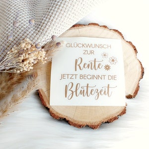 May include: A white card with gold lettering on a wooden slice. The card says "Gl&uuml;ckwunsch zur Rente Jetzt beginnt die Bl&uuml;tezeit" which translates to "Congratulations on your retirement Now the bloom time begins".