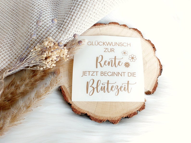 May include: A white card with gold lettering on a wooden slice. The card says "Gl&uuml;ckwunsch zur Rente Jetzt beginnt die Bl&uuml;tezeit" which translates to "Congratulations on your retirement Now the bloom time begins".