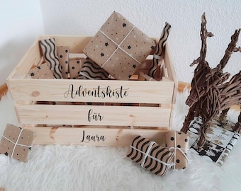 May include: A wooden crate with the words "Adventskiste für Laura" on the front. The crate is filled with small wrapped gifts, each wrapped in brown paper with black and white patterns.