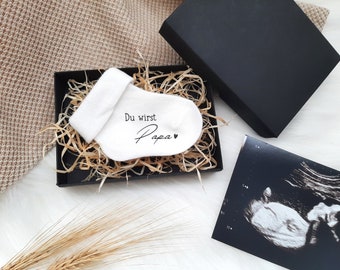 Könnte beinhalten: Eine weiße Baby-Socke mit dem Text "Du wirst Papa" und einem kleinen Herz darauf. Die Socke befindet sich in einer schwarzen Box mit braunem, zerkleinertem Papier. Ein Schwarzweiß-Ultraschallbild ist ebenfalls auf dem Foto zu sehen.