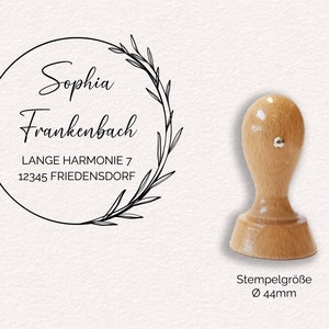 Könnte beinhalten: Ein selbsttinte­nder Stempel aus Holz mit einem schwarz-weißen Design eines Kranzes mit Blättern und dem Text "Sophia Frankenbach, Lange Harmonie 7, 12345 Friedersdorf". Der Stempel hat einen Durchmesser von 44 mm.