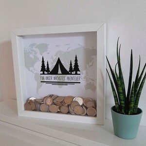 Peut inclure: Bo&icirc;te &agrave; ombre en bois blanc avec un fond de carte du monde et une silhouette noire d'une tente et d'arbres. Le texte "FUR UNSER NACHSTES ABENTEUER" est imprim&eacute; sous la tente. La bo&icirc;te est remplie de pi&egrave;ces de monnaie.