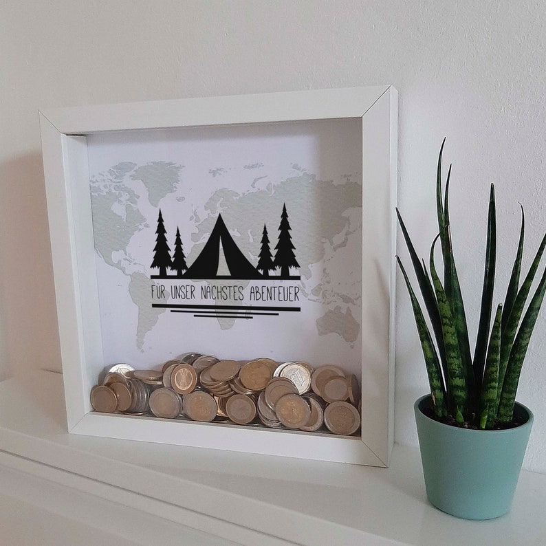 Peut inclure: Bo&icirc;te &agrave; ombre en bois blanc avec un fond de carte du monde et une silhouette noire d'une tente et d'arbres. Le texte "FUR UNSER NACHSTES ABENTEUER" est imprim&eacute; sous la tente. La bo&icirc;te est remplie de pi&egrave;ces de monnaie.