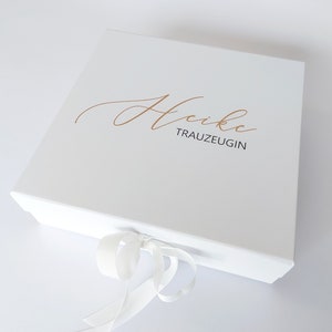 Könnte beinhalten: Weiße Geschenkbox mit einem weißen Satinband. Die Box trägt den Namen "Heike" in goldener Kursivschrift und das Wort "Trauzeugin" in Gold unter dem Namen.