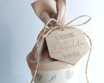 May include: A beige felt cone-shaped gift bag tied with twine, featuring a wooden tag. The tag is engraved with the word "SCHULKIND", the name "Mathilda", and the date "13.09.2021". The bag has a light beige felt top.