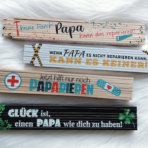 Könnte beinhalten: Vier Holz-Klapplineale mit verschiedenen Sprüchen. Das oberste Lineal ist rot, weiß und blau mit dem Text "keine Panik! Papa kann das reparieren!" Das zweite Lineal ist weiß mit gelben und schwarzen Streifen und dem Text "WENN PAPA ES NICHT REPARIEREN KANN, KANN ES KEINER!" Das dritte Lineal ist braun mit einem roten Kreuz und dem Text "Jetzt hilft nur noch PAPARIEREN". Das unterste Lineal ist schwarz mit grünen Kleeblättern und dem Text "GLÜCK ist, einen PAPA wie dich zu haben!"