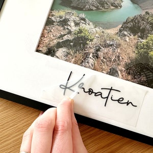 Könnte beinhalten: Ein weißer Bilderrahmen mit schwarzem Rand, ein Foto eines Flusses und Bergen und ein transparenter Aufkleber mit dem Wort "Kroatien" in schwarzer Kursivschrift.