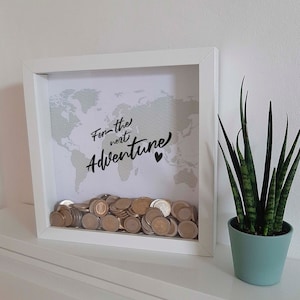 Peut inclure: Bo&icirc;te &agrave; ombre en bois blanc avec un fond de carte du monde et le texte "For the next Adventure" en &eacute;criture noire. La bo&icirc;te est remplie de pi&egrave;ces de monnaie.