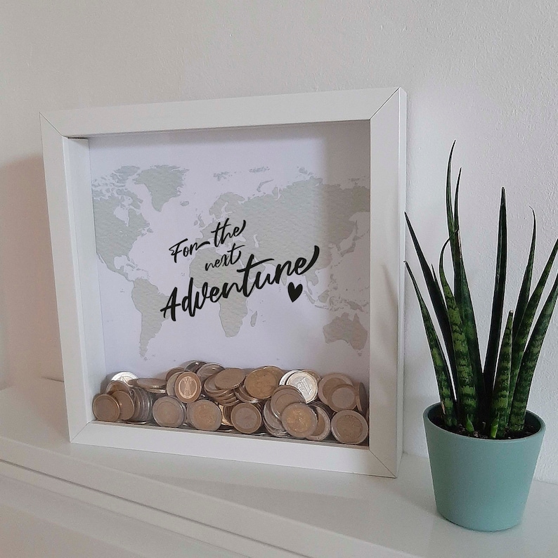 Peut inclure: Bo&icirc;te &agrave; ombre en bois blanc avec un fond de carte du monde et le texte "For the next Adventure" en &eacute;criture noire. La bo&icirc;te est remplie de pi&egrave;ces de monnaie.