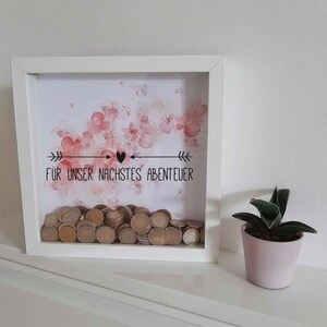 Peut inclure: Bo&icirc;te en bois blanche avec un motif floral aquarelle rose et le texte "F&Uuml;R UNSER N&Auml;CHSTES ABENTEUER" avec un symbole de c&oelig;ur. La bo&icirc;te est remplie de pi&egrave;ces de monnaie.