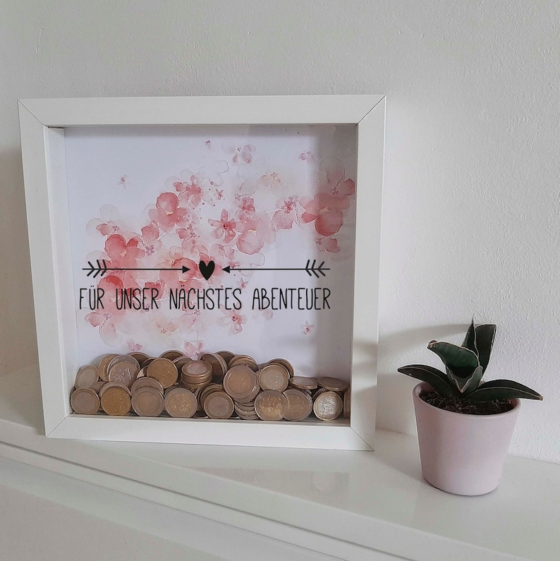 Peut inclure: Bo&icirc;te en bois blanche avec un motif floral aquarelle rose et le texte "F&Uuml;R UNSER N&Auml;CHSTES ABENTEUER" avec un symbole de c&oelig;ur. La bo&icirc;te est remplie de pi&egrave;ces de monnaie.