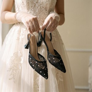 Elegant lace slingback heels with pointed toe and sculpted heel, available in white and black colors, styled in romantic bridal setting with pearls, lace fabric, and floral details for wedding or special occasion wear.
