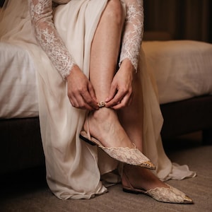 May include: A person is adjusting the ankle strap of a pair of beige lace flats with gold-colored toe caps. The flats are paired with a long, flowing cream-colored dress. The sleeves of the dress are made of lace.