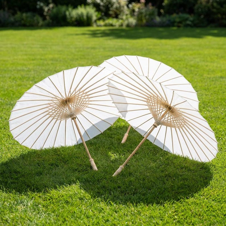 May include: Three white paper parasols with bamboo frames are arranged on a green lawn. The parasols are open, displaying their delicate, semi-transparent design. The handles are made of light-colored wood.
