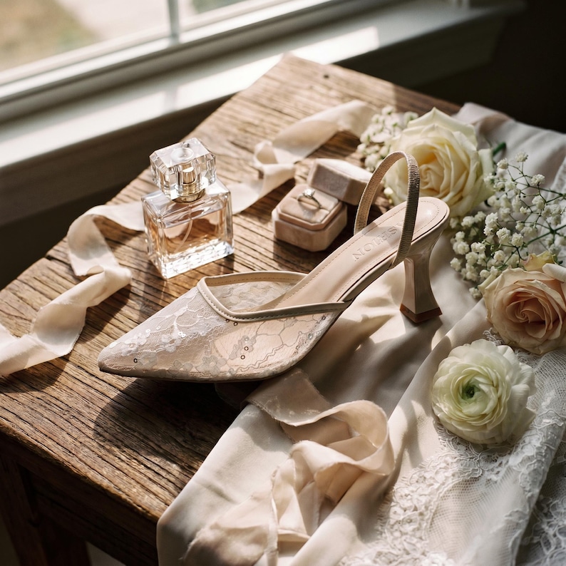 Elegant lace slingback heels with pointed toe and sculpted heel, available in white and black colors, styled in romantic bridal setting with pearls, lace fabric, and floral details for wedding or special occasion wear.