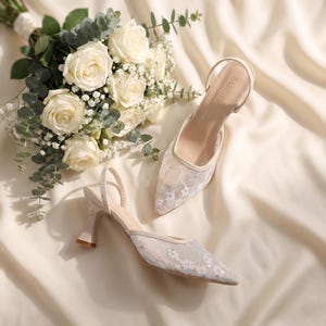 Elegant lace slingback heels with pointed toe and sculpted heel, available in white and black colors, styled in romantic bridal setting with pearls, lace fabric, and floral details for wedding or special occasion wear.