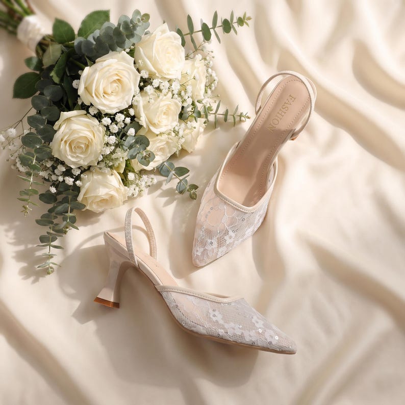 Elegant lace slingback heels with pointed toe and sculpted heel, available in white and black colors, styled in romantic bridal setting with pearls, lace fabric, and floral details for wedding or special occasion wear.