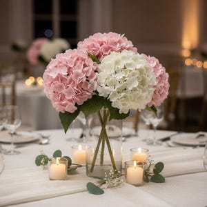 May include: A floral centrepiece with pink and white hydrangeas in a clear glass vase. The arrangement is on a white tablecloth, with lit candles in glass holders. Green leaves and small white flowers add to the decor, creating a soft, romantic ambiance.