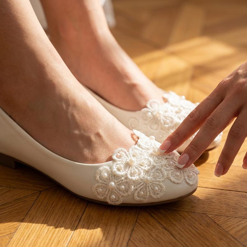 Ivory bridal lace flats with pearl details on feet, elegant wedding ballet shoes with floral lace design, comfortable bride shoes for ceremony and reception on wooden floor