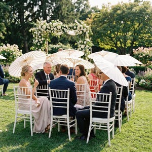 May include: Outdoor wedding reception with guests seated at white chairs, holding white paper parasols. A floral archway and lush greenery create a picturesque setting. People are dressed in formal attire, enjoying the event.