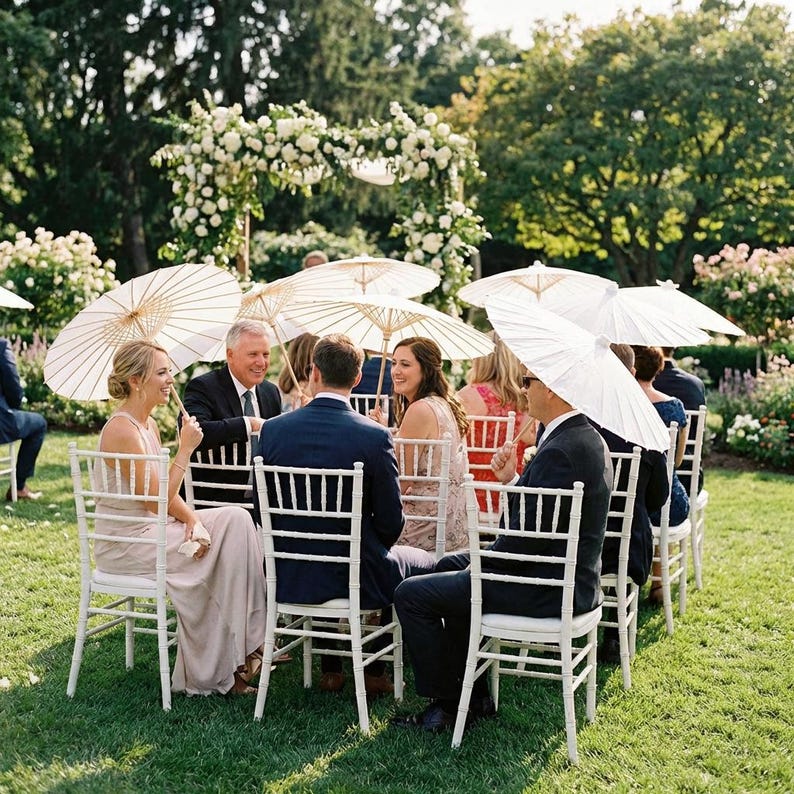May include: Outdoor wedding reception with guests seated at white chairs, holding white paper parasols. A floral archway and lush greenery create a picturesque setting. People are dressed in formal attire, enjoying the event.