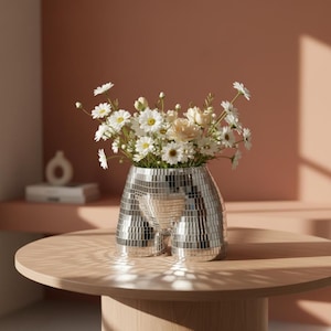 Könnte beinhalten: Eine silberne Mosaikvase in Form eines Torso, gefüllt mit weißen und gelben Blumen. Die Vase steht auf einem runden Holztisch. Der Hintergrund ist ein gedämpftes Rosa. Warmes Licht beleuchtet die Szene.