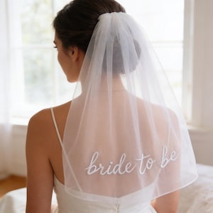 Op de afbeelding: Een doorschijnende witte bruidssluier met de geborduurde tekst "bride to be" in sierlijke letters. De sluier is gedrapeerd over de rug van een persoon die een witte jurk draagt. De achtergrond toont een raam en zacht licht.