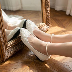 Ivory bridal lace flats with pearl details on feet, elegant wedding ballet shoes with floral lace design, comfortable bride shoes for ceremony and reception on wooden floor