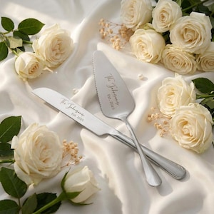Może przedstawiać: Srebrny zestaw noża i łopatki do ciasta z napisem "The Johnsons EST. 2025". Zestaw jest prezentowany na białej satynowej powierzchni, otoczony białymi różami i zielenią.