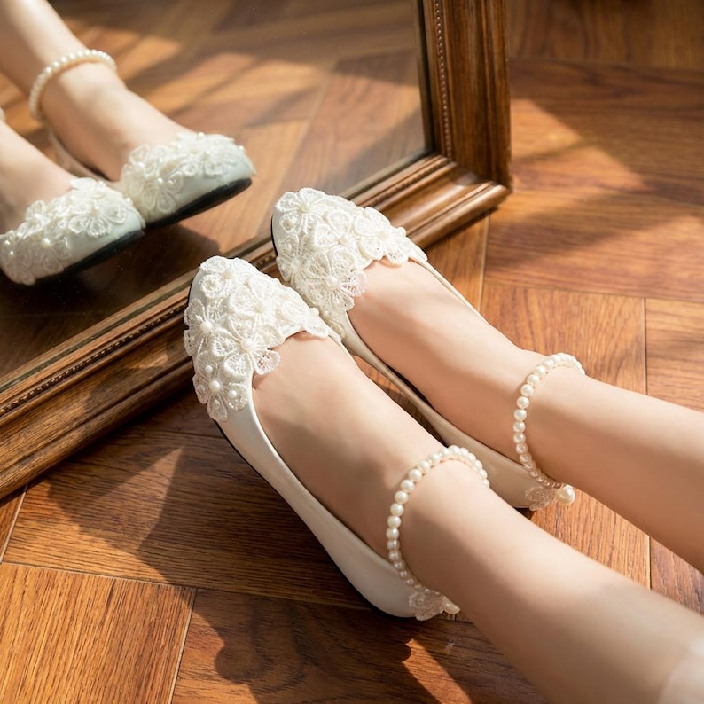 Ivory bridal lace flats with pearl details on feet, elegant wedding ballet shoes with floral lace design, comfortable bride shoes for ceremony and reception on wooden floor