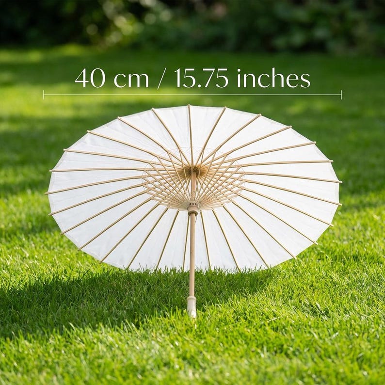 May include: A white paper parasol with a wooden handle and ribs, resting on green grass. The parasol is open, displaying its circular shape. The text above the parasol reads "40 cm / 15.75 inches".
